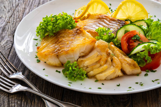 istock Fried codfish loin and fresh vegetables served on white plate on wooden table. 2190189290