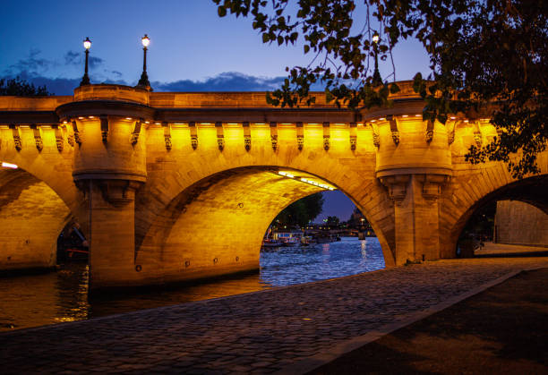Illuminated old bridge arch on the Seine River in Paris seen from the pier under the blue sky at dusk in Paris, France