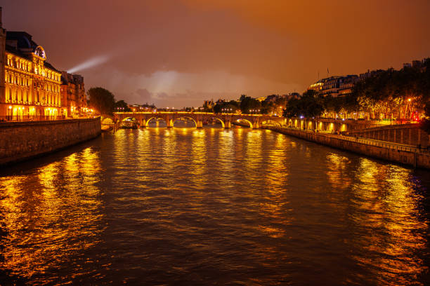 Illuminated old arch bridge and riverbanks of the Seine River in Paris under the cloudy bronze sky at night in Paris, France