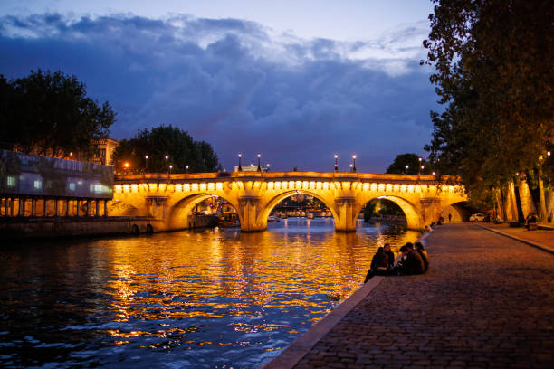 Paris, France, June 7, 2022: People on the promenade by the Seine River, illuminated bridge Pont Neuf over the river after sunset, Paris, France