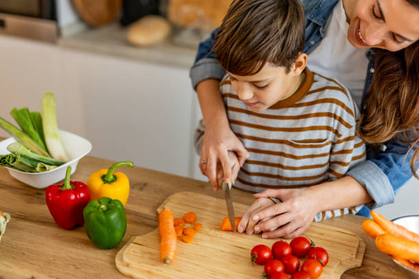 istock Mother carefully assisting her son while he's learning to cut carrots on a wooden board, surrounded by fresh vegetables 2190213451