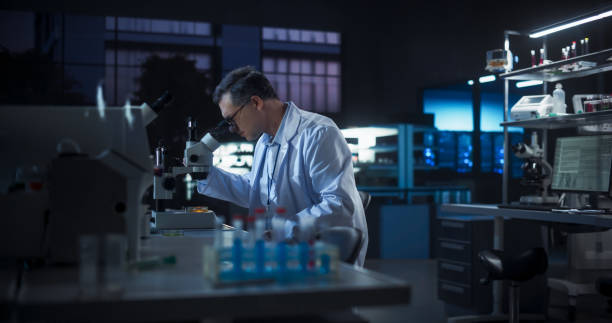 istock Molecular Biology Research Scientist Looking at a DNA Sample Under a Microscope in an Applied Science Laboratory. Portrait of an Adult Lab Engineer in White Coat Working on Vaccine Late at Night 2190228081