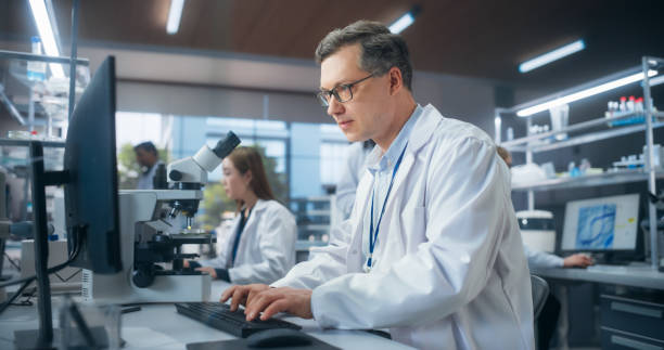 istock Handsome Adult Scientist Wearing Glasses and a White Lab Coat, Studying DNA Samples and Evaluating Data on Computer. Group of Multiethnic Specialists Working in a Laboratory 2190229173