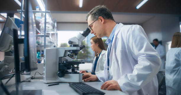 istock Excited Motivated Biotechnologist Studying Genetic Samples Under a Microscope and Researching Data on Computer in Order to Develop New Medical Products. Group of Scientists Working in a Laboratory 2190229250