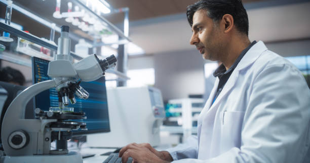 istock Genetic Research and Development Laboratory: Adult Scientist Working on a Computer and Looking at a Sample in a Petri Dish Under a Microscope. Advanced Biotechnology Lab with Modern Equipment 2190229410