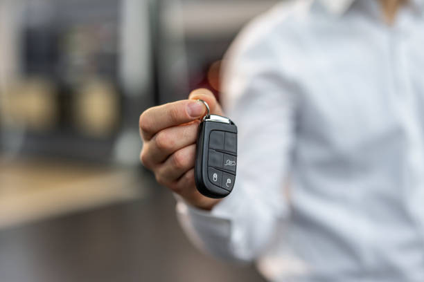 A young, smiling sales advisor at a car dealership holds out the keys to the vehicle, symbolizing the moment when dreams become reality. His friendly expression and professional appearance in a white shirt create an atmosphere of trust and satisfaction.