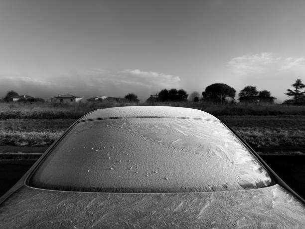 A black and white image capturing a car covered in frost on a cold morning. The intricate frost patterns are visible on the rear windshield and roof, glistening faintly. The background features a field and distant houses under a clear sky, emphasizing the chilly atmosphere.