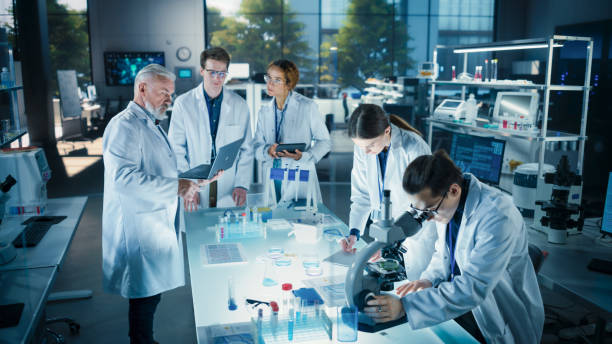 istock Head of Laboratory Making a Bioengineering Experiment with a Group of Young Science College Students. Future Specialists Working with Modern Microscopes, Using Tablet and Laptop Computers 2190243603