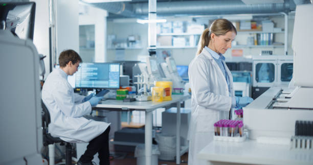 istock Biomedical Researchers Conduct Experiments in a Modern Laboratory. Female Scientist Preparing a Set of Samples in Test Tubes with Purple Caps. Colleague Analyzes Data on a Computer in the Background 2190246669