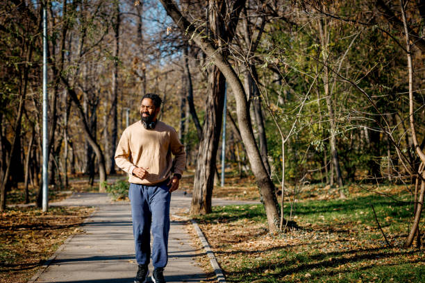 Young adult man wearing casual clothes is jogging in a park during a sunny autumn day, enjoying the fresh air and the warm sunlight while listening to music