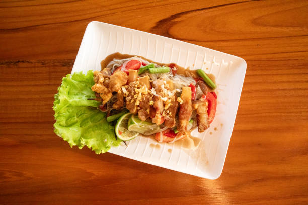 Small noodle salad with fermented fish and crispy pork with vegetables and tomatoes on the side Thai style long beans and chilli on a white plate