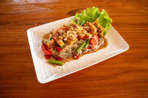 Small noodle salad with fermented fish and crispy pork with vegetables and tomatoes on the side Thai style long beans and chilli on a white plate