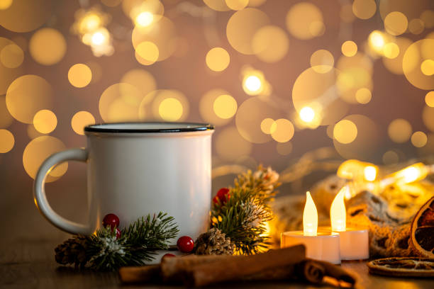Front view of a hot white cup surrounded by cinnamon sticks, dried orange slices and some Christmas decoration. At the background are some defocused Christmas lights and can be used as a useful copy space