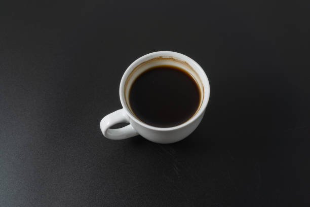 Detailed close-up from above of an old-fashioned white porcelain mug filled with freshly poured black coffee, with a bit of foam on top. the background is white with a soft shadow gradient to the right. the handle is on the left