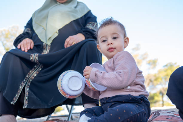 A young child sitting on a mat outdoors, holding bowls, with a family member nearby. The environment is sunny and serene, portraying warmth and familial moments of play and connection
