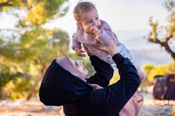 A joyful mother in a headscarf lifts her smiling baby above her head in a sunlit garden. The scene captures a moment of love and happiness in a natural setting.