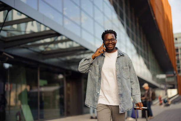 istock Man Joyfully Walking with Luggage and Talking on Phone near Modern Building 2190270123