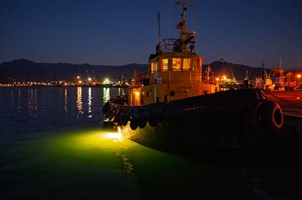 Tugboat illuminated by dock lights at night, with vibrant reflections in the water.