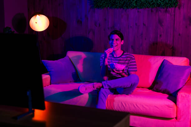 istock Young man enjoying a relaxed evening eating popcorn while watching television in a cozy, vividly lit living room 2190279248