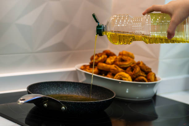 A person is pouring olive oil into the pan from a plastic bottle. He is in the kitchen. The pan is on the stove. In the background you can see a cooking dish filled to the top with a pile of freshly made donuts.