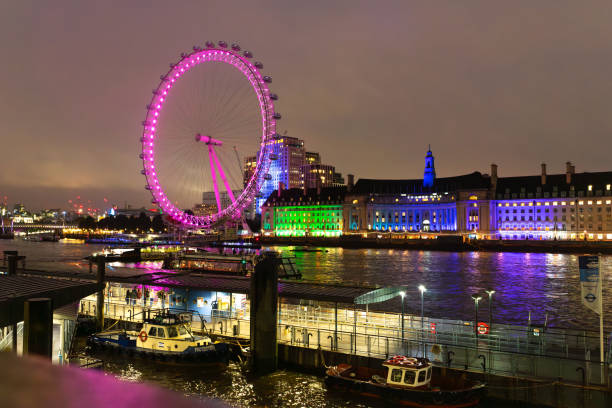 The London Eye illuminated in vibrant pink lights at night, reflecting off the River Thames with the surrounding cityscape and buildings glowing in blue and green hues.