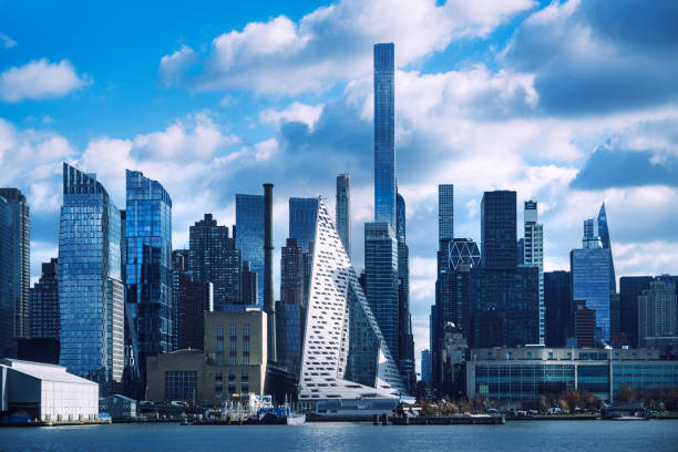 A contemporary city skyline showcasing diverse architectural designs reflecting in the calm river, under a partly cloudy sky, capturing the essence of urban development in New York City.