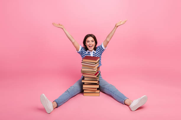 istock Young woman with happy expression and raised arms sitting on pink background with a stack of books, wearing a striped casual outfit 2190285826