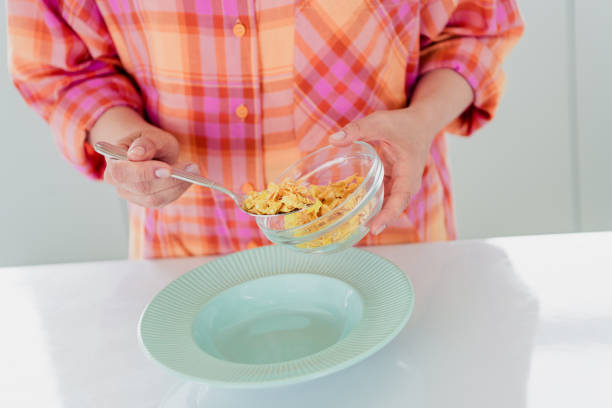 Mature woman in casual attire pouring cereal into a bowl in a well-lit kitchen, reflecting comfort and culinary delight.