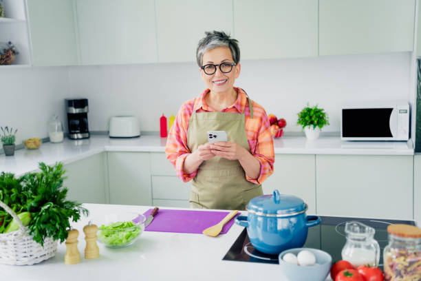 istock Elderly woman cooking in modern kitchen, smiling happily while using phone 2190288796