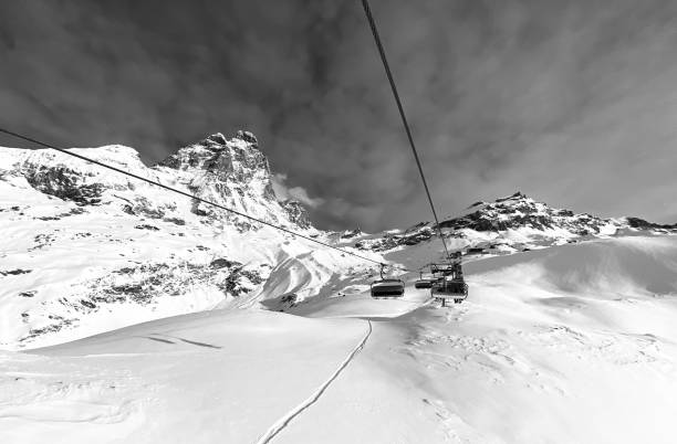 The photo depicts a stunning winter landscape in black and white, featuring a ski lift with empty chairs suspended above a snowy expanse. In the background, a majestic mountain rises sharply against a cloudy sky, its peaks capped with snow. The foreground shows a smooth, untouched blanket of snow, with a single ski track leading towards the mountains, highlighting the serene and pristine nature of the alpine environment.