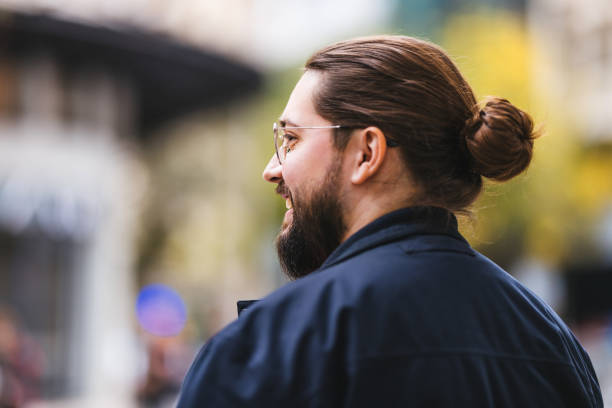 A stylish young man sporting a beard and wearing glasses is smiling while standing outdoors on a city street with a blurred background, exuding warmth and authenticity.