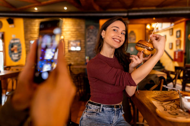 In a bustling restaurant, a young woman smilingly poses with a mouthwatering burger, eagerly waiting for her friend to snap the perfect photo to share on social media