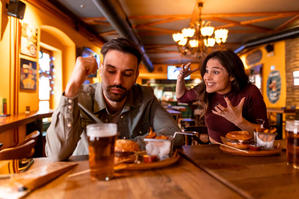 Young woman teasing bored man at restaurant table Young woman making funny faces and playfully teasing a bored young man while enjoying hamburgers and fries at a lively restaurant table, creating a cheerful atmosphere brat burger stock pictures, royalty-free photos & images