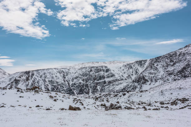 istock Cold snowy alpine landscape with last year's grass, sharp rocks and stones in white snow against snow-covered rocky mountain range under clouds in blue sky. High snow-white mountains in sunny day. 2190303968