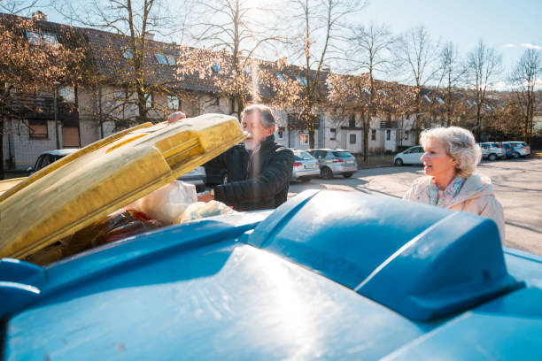An elderly man and woman deposit recyclables into a blue bin, demonstrating their commitment to environmental sustainability in a sunny outdoor setting.