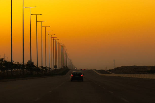 The image depicts a multi-lane highway stretching towards the horizon under a vibrant, intensely orange sunset. A line of tall, slender light poles runs parallel to the road on the left, creating a strong leading line that draws the eye towards the vanishing point. Several cars are visible, traveling in the same direction, appearing as dark silhouettes against the bright background. The overall atmosphere is serene yet somewhat lonely, with the warm colors of the sunset contrasting with the dark road and vehicles. The air appears hazy, softening the details in the distance. The scene suggests a feeling of vastness and open space.