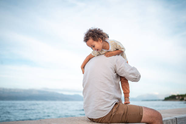 A Caucasian father and his diverse young son playfully engage on a seaside pier. The father, dressed in casual shorts and shirt, lifts his son, who wears a striped shirt and pants, creating a joyful family moment by the ocean.