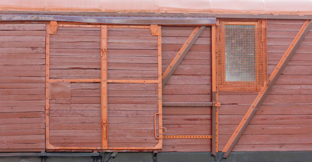 The red door of a building has a window on the side. The door is old and rusted, and the window is small and has a metal frame. Scene is somewhat nostalgic and somewhat eerie, as the old