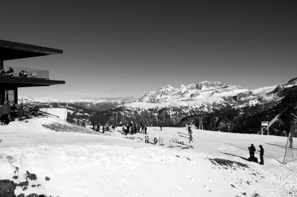 This black and white photograph depicts a mountain lodge situated in the Dolomites, surrounded by a stunning snowy landscape. The image showcases the modern structure of the lodge on the left, with people relaxing on its terrace and others enjoying outdoor winter activities. In the background, the majestic snow-capped peaks of the Dolomites dominate the horizon, while skiers and visitors scatter across the open slopes. The serene, crisp atmosphere and the contrast between the lodge and the dramatic mountain scenery highlight the beauty of this winter paradise