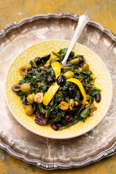 Still life food photography. Bakkoula, Moroccan olive and spinach salad on yellow ceramic plate with silver plate background.