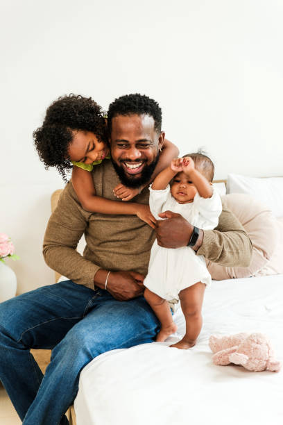 istock A joyful father sits on a bed, playfully holding his baby while his daughter hugs him, creating a heartwarming family moment. Happy diversity family concept. 2190354539