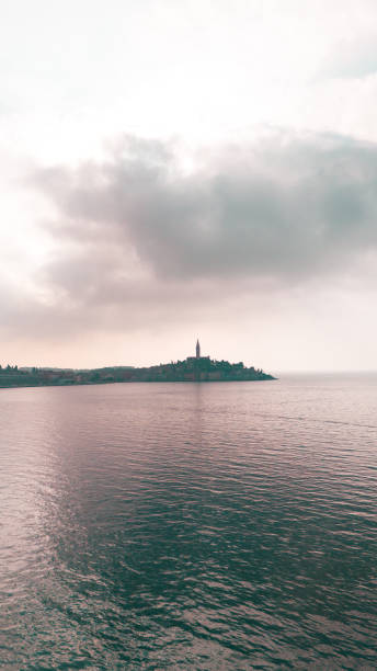 A vertical view of Rovinj, Croatia, featuring the iconic silhouette of the historic town and its church tower under a dramatic, cloudy sky. The calm Adriatic Sea reflects the muted tones of the scene, evoking a sense of serenity and timeless coastal charm.