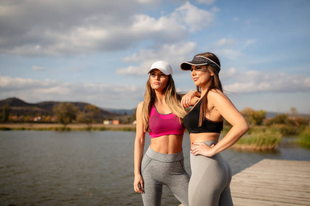 Active and healthy women posing outdoors in athletic wear, radiating confidence and joy by a tranquil lakeside backdrop with clear skies.