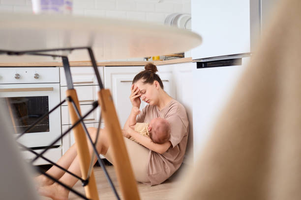 A tired mother sits on the kitchen floor with her newborn, resting her head on her hand in a moment of stress. The bright kitchen environment adds to the feeling of exhaustion in her gaze