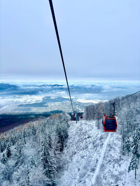 A picturesque cable car ride over a snow-covered forest with breathtaking views of the winter landscape and distant town below. The red gondola contrasts against the white snow and misty blue sky, creating a serene and scenic winter travel experience.