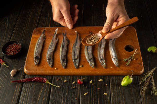 Hands skillfully arrange fresh fish on a rustic cutting board, sprinkling aromatic herbs and spices. A warm kitchen ambiance enhances the culinary artistry and anticipation of a delicious meal.