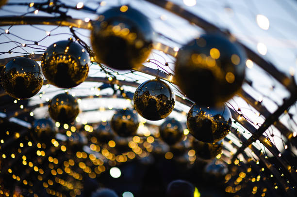 Close-up of golden Christmas ornaments hanging from a tree with glowing festive lights in the background. The ornaments reflect the warm holiday atmosphere.