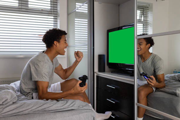 Medium side-view shot of a teenage boy sitting on the edge of his bed playing video games at home in Sunderland, North East England. He is holding a games controller, raising one fist in celebration towards the screen. The screen is Chroma Key colour.Videos similar to this scenario available.