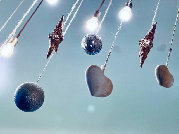 Showcasing delicate hanging ornaments, including glittery spheres, heart shapes, and small textured decorations suspended by strings under soft glowing lights. The muted blue background complements the festive and dreamy atmosphere.