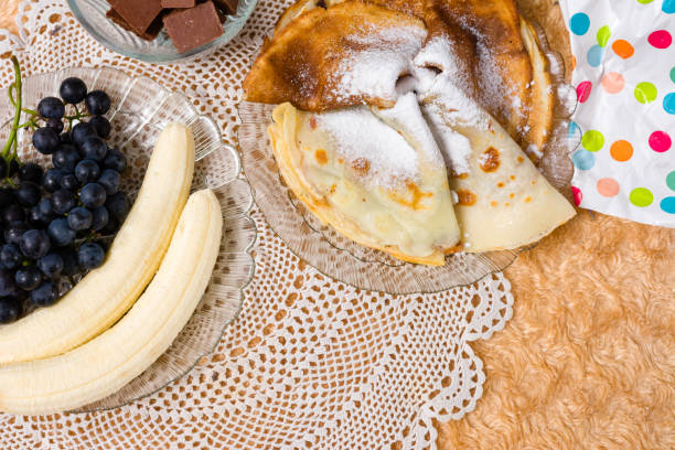 Thin pancakes dusted with powdered sugar are presented on a delicate doily alongside fresh bananas, grapes, and chocolate pieces, creating a tempting dessert spread.